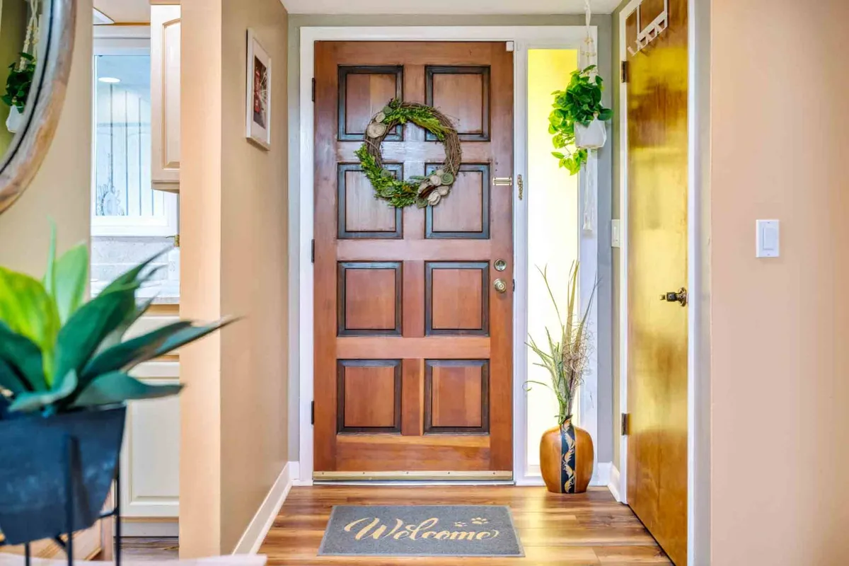 An interior shot of the home's entryway, featuring a wooden front door adorned with a wreath, a welcoming mat, and green plants. The image conveys a warm, inviting atmosphere that sets the tone for a comfortable stay.