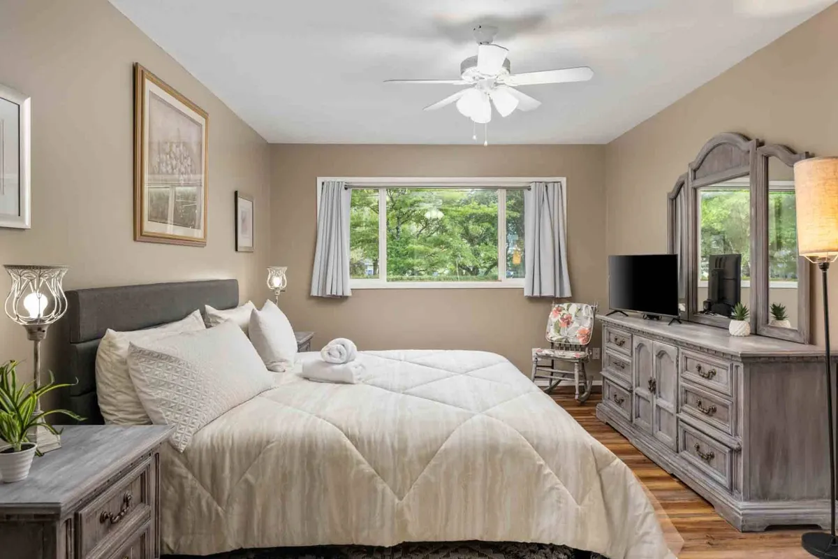 A tranquil bedroom highlighted by a bed adorned with decorative pillows, a window that floods the room with sunlight, a nightstand with a lamp for evening reading, and a dresser with a mirror. The subtle, soothing color palette enhances the peaceful retreat feel.