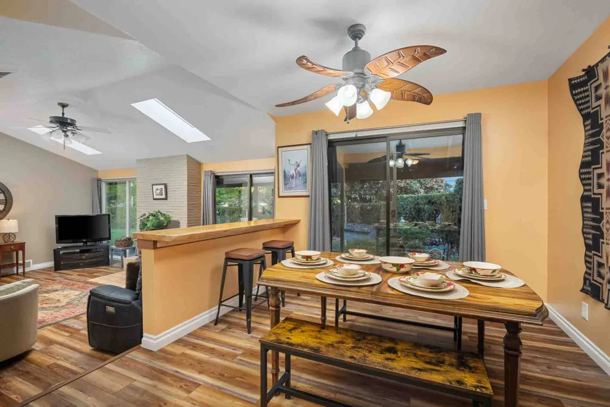 A warm and inviting dining area with a wooden dining table set with plates, a spiral ceiling fan above, and large glass doors that open to an outdoor space. The area seamlessly connects to the living space, emphasizing the open-concept layout.