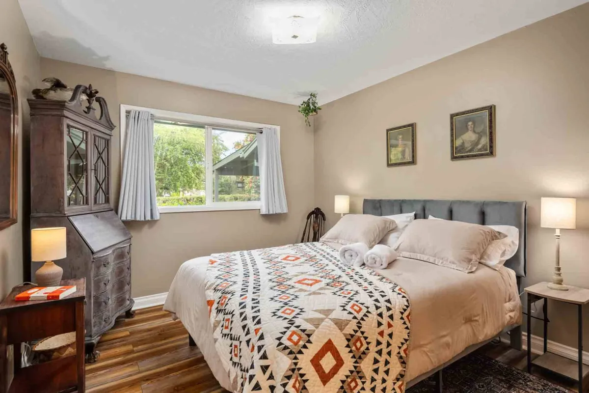 A cozy bedroom featuring a comfortable bed with soft linens, a wardrobe for personal storage, a large window letting in natural light, decorative wall art, and bedside lamps for reading or ambiance. The calming décor and ample space create a restful atmosphere.