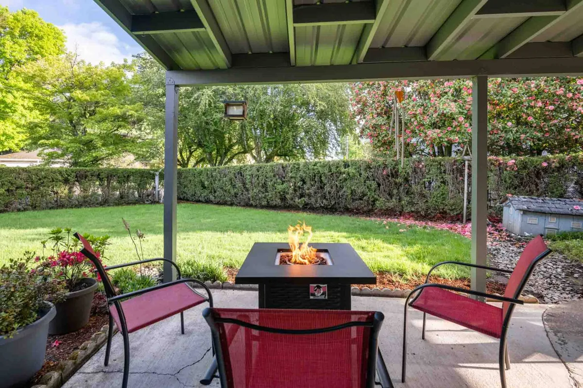 This inviting exterior shot features a fire pit with red chairs set amidst lush greenery and a garden hedge. The intimate setup is perfect for family gatherings, evening relaxation, and quiet mornings with coffee. The greenery and fencing suggest a secure and private backyard.