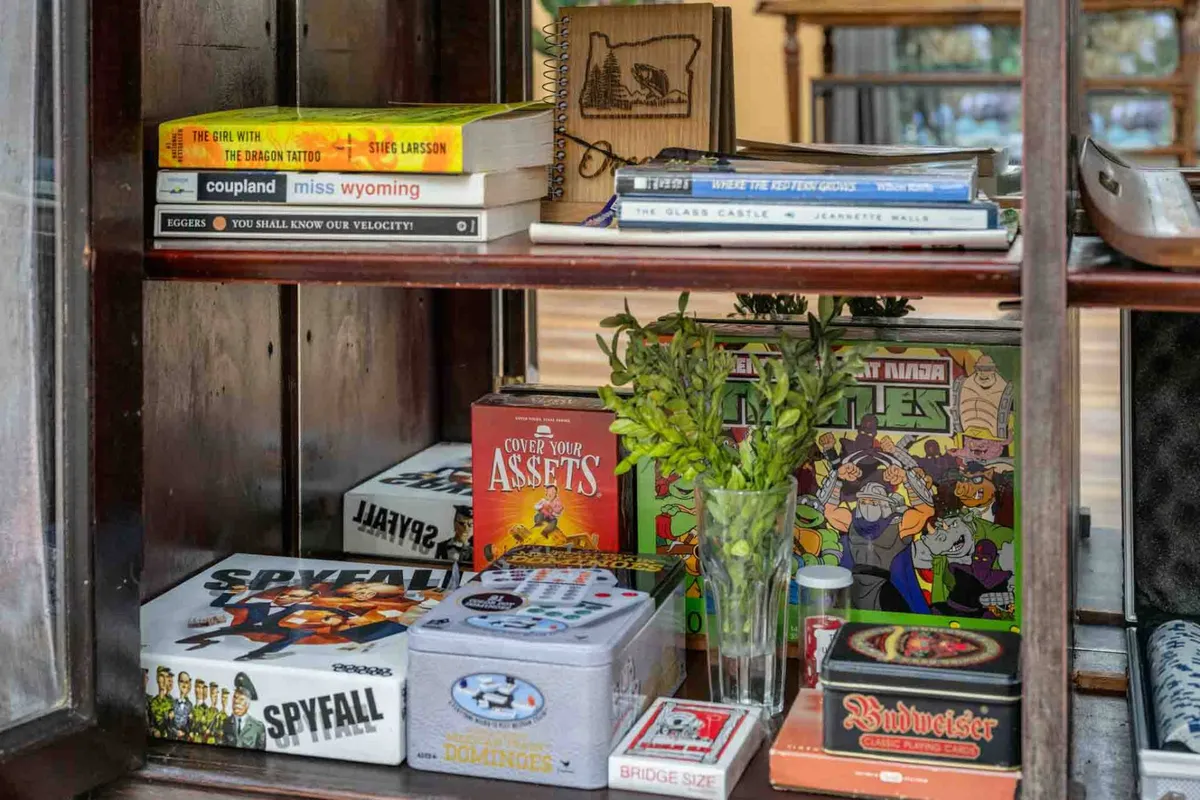 A living area featuring a shelf filled with books and board games, a vase with greenery, and mixed media decor. The scene is cozy and engaging, suggesting a space where families and friends can gather for board games, reading, and casual socializing.