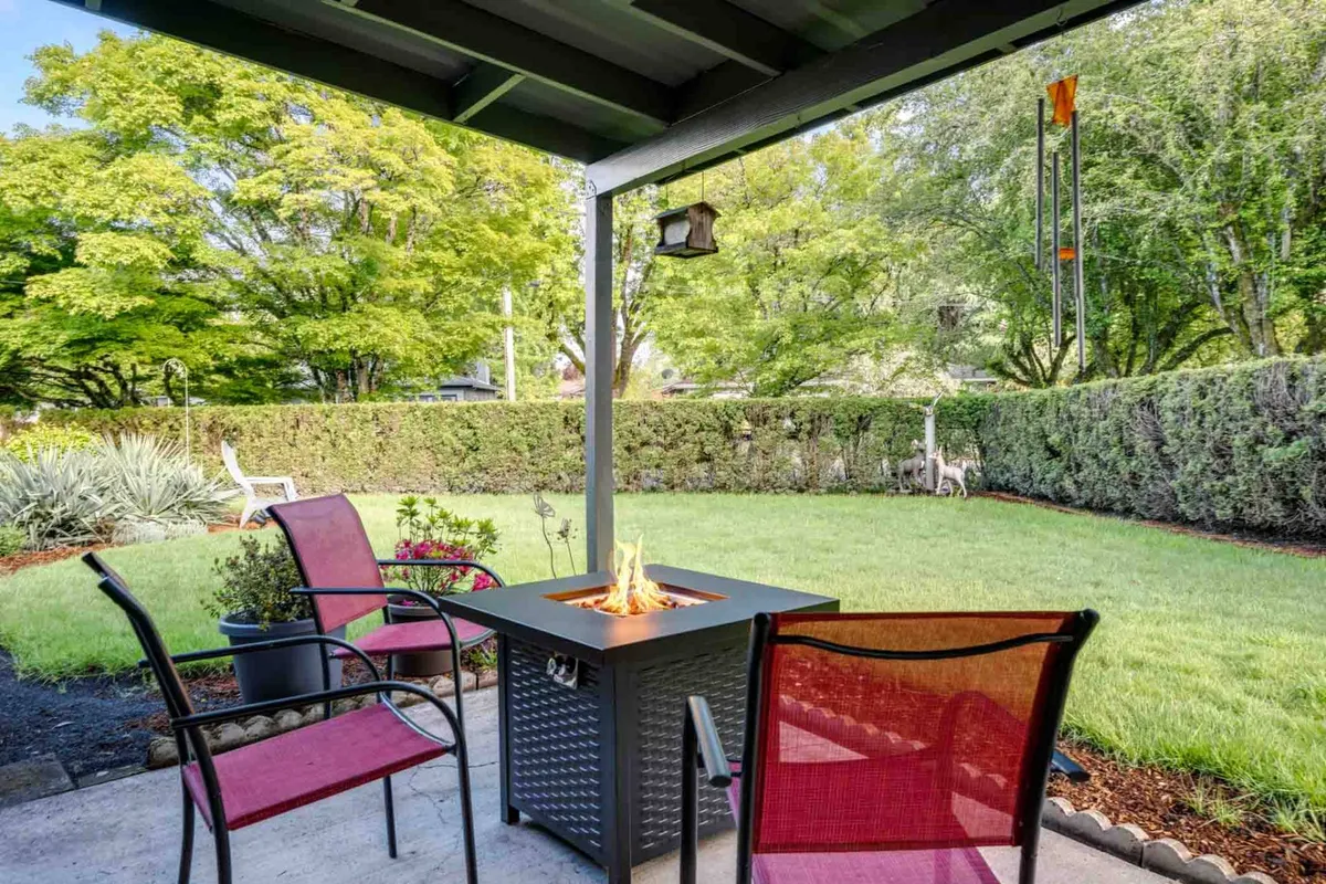 This scene shows a cozy, covered outdoor seating area with a fire pit, lush greenery, and wind chimes. The mood is tranquil and serene, ideal for relaxing, socializing, or enjoying nature in a peaceful setting. The covered aspect and fire feature invite both daytime and evening use.
