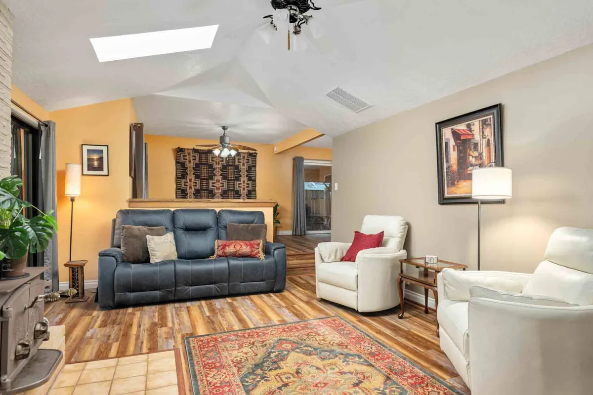 A cozy living room furnished with a comfortable gray sofa, white armchairs, a colorful area rug, and decorative wall art. The space is well-lit with natural light, and modern lighting fixtures add warmth. The setup is ideal for socializing, reading, or hosting lively gatherings.