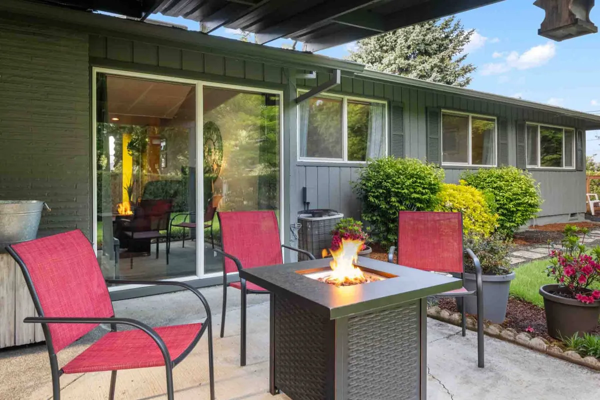 This exterior image captures a cozy backyard patio area featuring a fire pit surrounded by red chairs, lush greenery, and a sliding glass door leading into the home. The setting evokes a welcoming space for relaxation or socializing after a busy day.