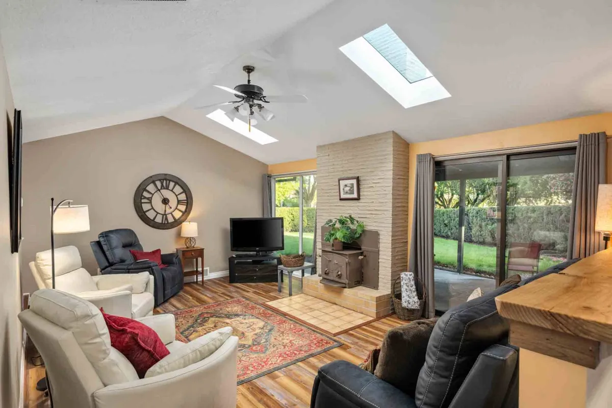 The image showcases a cozy living room with a plush sofa set arranged around a contemporary coffee table, facing a large television and a classic fireplace. Natural light streams in through overhead skylights, creating a warm and inviting atmosphere ideal for relaxation or gathering with loved ones.